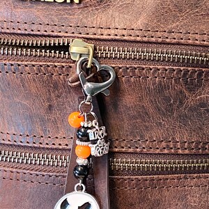 May include: A brown leather bag with a zipper and a charm attached to the zipper pull. The charm features a black and white Jack Skellington face, orange and black beads, and a silver charm that says "Trick or Treat".