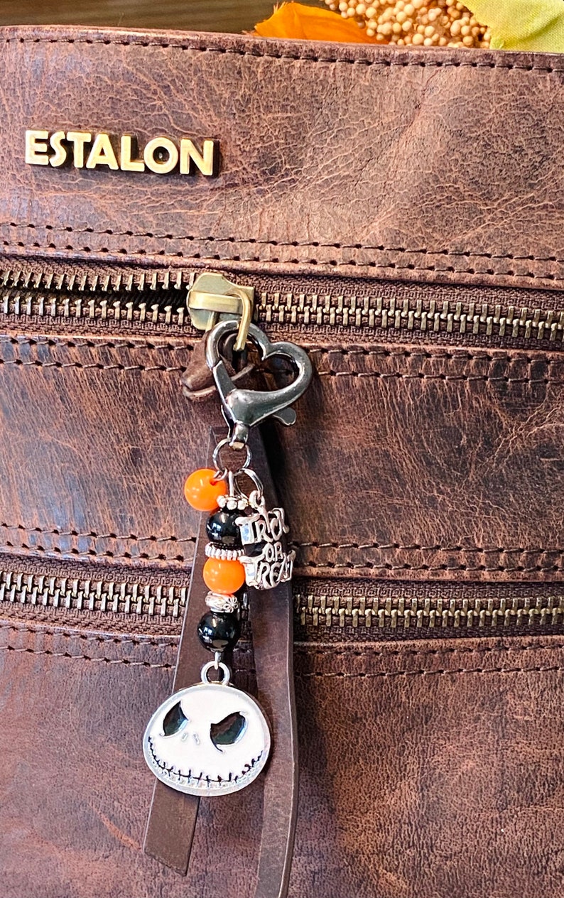 May include: A brown leather bag with a zipper and a charm attached to the zipper pull. The charm features a black and white Jack Skellington face, orange and black beads, and a silver charm that says "Trick or Treat".