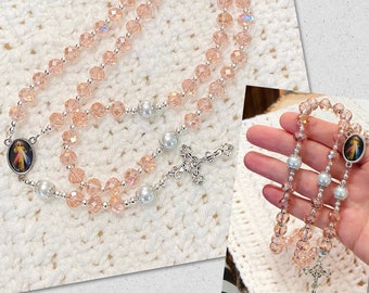 May include: A rosary featuring peach-colored faceted beads and silver-toned accents. The rosary includes a crucifix, a centerpiece with an image, and faux pearl beads. The beads are strung together with silver-colored wire.