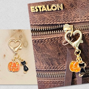 May include: Gold-tone pumpkin and black cat charm with a heart-shaped clasp. The charm is attached to a brown leather bag with the word "ESTALON" embossed on the front.