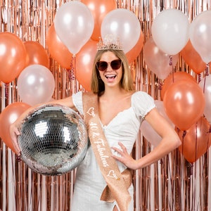 May include: A woman wearing a white dress and a gold sash with the text "40 & Fabulous" is holding a silver disco ball. She is wearing a tiara and sunglasses. The background is a wall of rose gold foil.