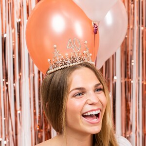 May include: A woman wearing a gold tiara with rhinestones and pearls, the tiara has the number 40 on it. She is smiling and looking to the right. The background is a rose gold foil curtain.
