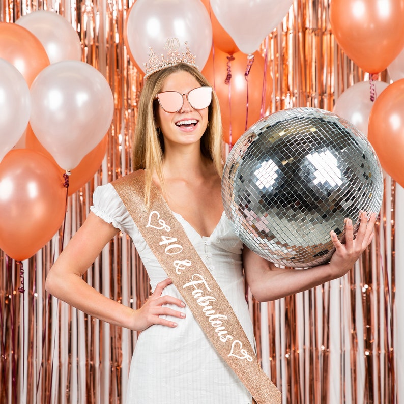 May include: A woman wearing a white dress and a gold glitter sash that says "40 & Fabulous" holds a silver disco ball in front of a rose gold backdrop with balloons.