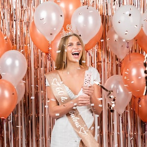 Op de afbeelding: Een vrouw in een witte jurk en een gouden sjerp met de tekst "60 & Fabulous" met een kroon op haar hoofd. Ze houdt een confettikanon vast en glimlacht. Er zijn roze en witte ballonnen en confetti op de achtergrond.