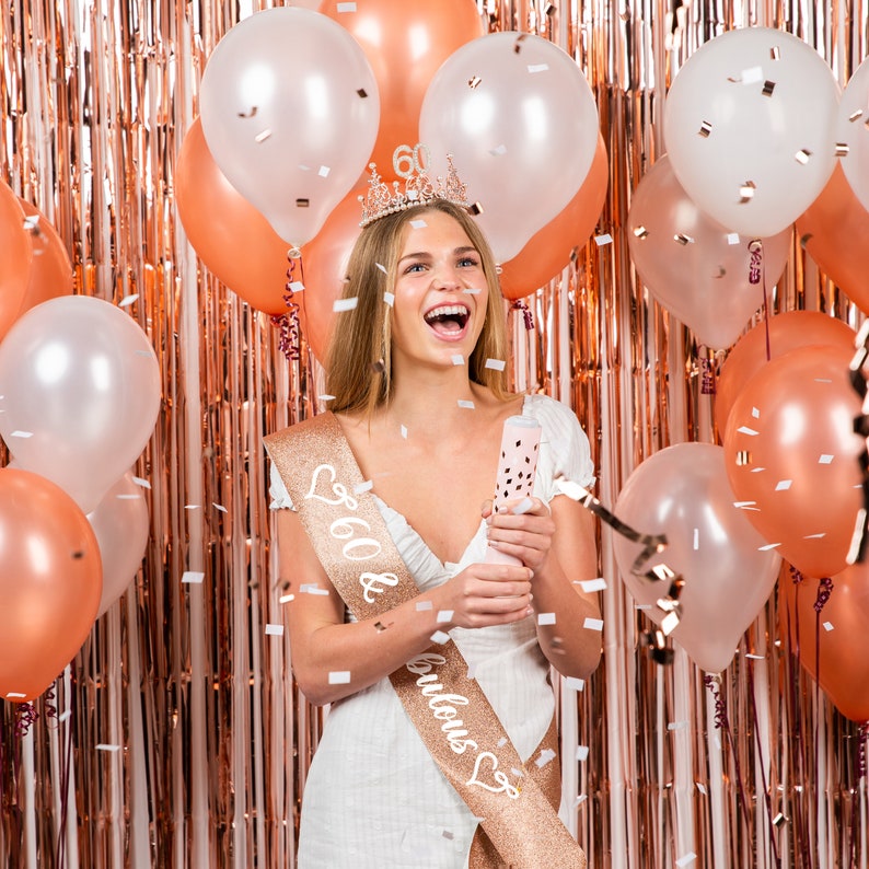 Op de afbeelding: Een vrouw in een witte jurk en een gouden sjerp met de tekst "60 & Fabulous" met een kroon op haar hoofd. Ze houdt een confettikanon vast en glimlacht. Er zijn roze en witte ballonnen en confetti op de achtergrond.