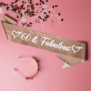 Op de afbeelding: Een gouden glitterband met de tekst "60 & Fabulous" in witte letters en een hartvormig ontwerp aan beide uiteinden. Een gouden hoofdband met parel accenten is ook afgebeeld.