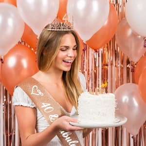 May include: A woman wearing a gold crown with the number 40 on it, a gold sash with the text "40 & fabulous" and a white shirt, holds a white cake with gold candles. The cake is on a white plate. The background is a rose gold foil curtain with balloons.