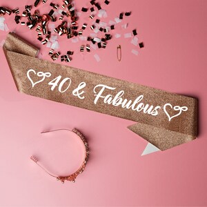 May include: A gold glitter sash with the text "40 & Fabulous" in white lettering and a heart design on each end. A gold headband with rhinestones is also pictured.