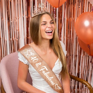 May include: A woman wearing a gold glitter sash with the text "40 & Fabulous" and a gold tiara with rhinestones. She is smiling and laughing.