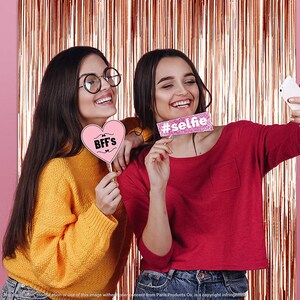 Op de afbeelding: Twee vriendinnen poseren voor een selfie voor een roze gouden tinsel achtergrond. Een vriendin houdt een roze hartvormig bord vast met de tekst "BFF's" en de andere houdt een roze bord vast met de tekst "#selfie".