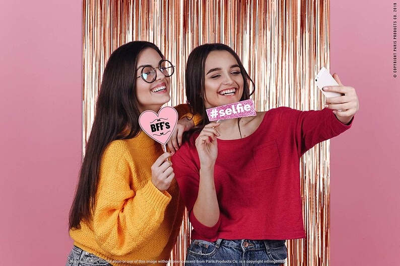 Op de afbeelding: Twee vriendinnen poseren voor een selfie voor een roze gouden tinsel achtergrond. Een vriendin houdt een roze hartvormig bord vast met de tekst "BFF's" en de andere houdt een roze bord vast met de tekst "#selfie".