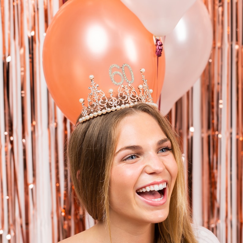 Op de afbeelding: Een vrouw draagt een gouden tiara met strass en parels, de tiara heeft het nummer 60 erop. Ze glimlacht en kijkt naar rechts. Er zijn twee ballonnen achter haar, een is perzikkleurig en de andere is wit.