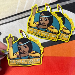 May include: Embroidered patches featuring a Native American woman with a stern expression and raised middle finger. The patches have text that reads "THE BAD ATTITUDE CLUB SINCE 1492" and "Land Back" in a yellow and teal color scheme.