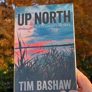 May include: A book titled "Up North: A North Country Mystery" by Tim Bashaw. The cover features a sunset over a lake with tall grass in the foreground.