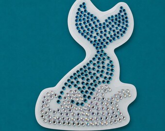 May include: A decorative patch shaped like a mermaid tail emerging from water, constructed with blue rhinestones forming the tail and clear iridescent rhinestones creating the wave base, all set on a white backing with a die-cut border.