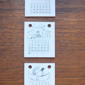 May include: Three white square calendar pages for January, February, and March 2027. Each page features black line art illustrations at the top, including orchids, a celebration scene with champagne, and a bird, with two holes at the top.