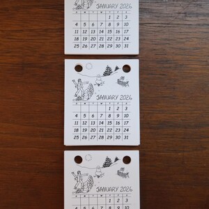 May include: Three small, white calendars for January 2026, each with a whimsical black and white illustration and calendar grid. The calendars have a hole at the top for hanging.
