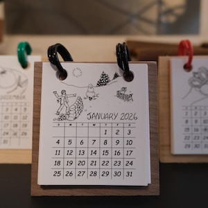 May include: A set of three small desk calendars with wooden bases. The calendars feature black and white illustrations and dates for December, January 2026, and March. Each calendar has a different colored loop at the top.