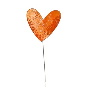 May include: An orange heart-shaped garden stake with a textured surface. The heart is mounted on a thin, dark metal rod. The stake is designed for outdoor use and adds a decorative touch to gardens.