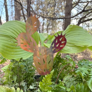 May include: A metal cactus garden stake with a copper finish. The cactus has a cut-out design and is surrounded by green foliage. The stake is designed to be placed in a garden or potted plant.