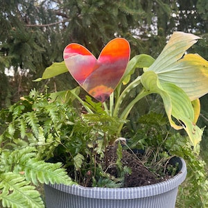 May include: A heart-shaped garden stake with a colorful sunset design is placed in a pot of green plants. The stake is made of metal and has a vibrant gradient of orange, red, and purple hues. The pot is gray with vertical white stripes.