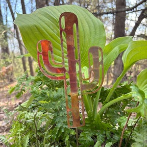 May include: A rusty-colored metal cactus garden stake. The cactus has a detailed cutout design and is placed in a garden with green plants and leaves. The stake is a decorative item for outdoor spaces.