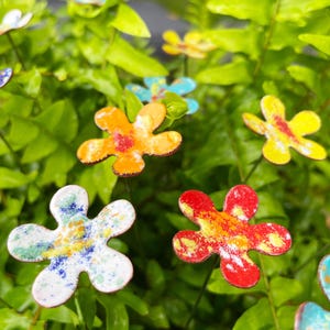 May include: Colorful metal flower garden stakes in various colors, including orange, yellow, red, and blue. The flowers have a speckled design and are set against a backdrop of green foliage. These decorative stakes add a touch of whimsy to any garden.