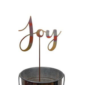 May include: A metal garden stake with the word "Joy" in a cursive font. The letters have a gradient of colors, including orange, yellow, and purple. The stake is inserted into a galvanized metal bucket.