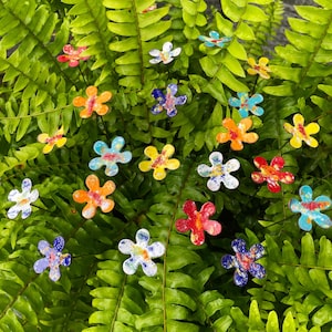 May include: A collection of colorful, flower-shaped garden stakes in various colors, including red, orange, yellow, blue, and white. The stakes are placed among green fern leaves, creating a vibrant and decorative garden accent.