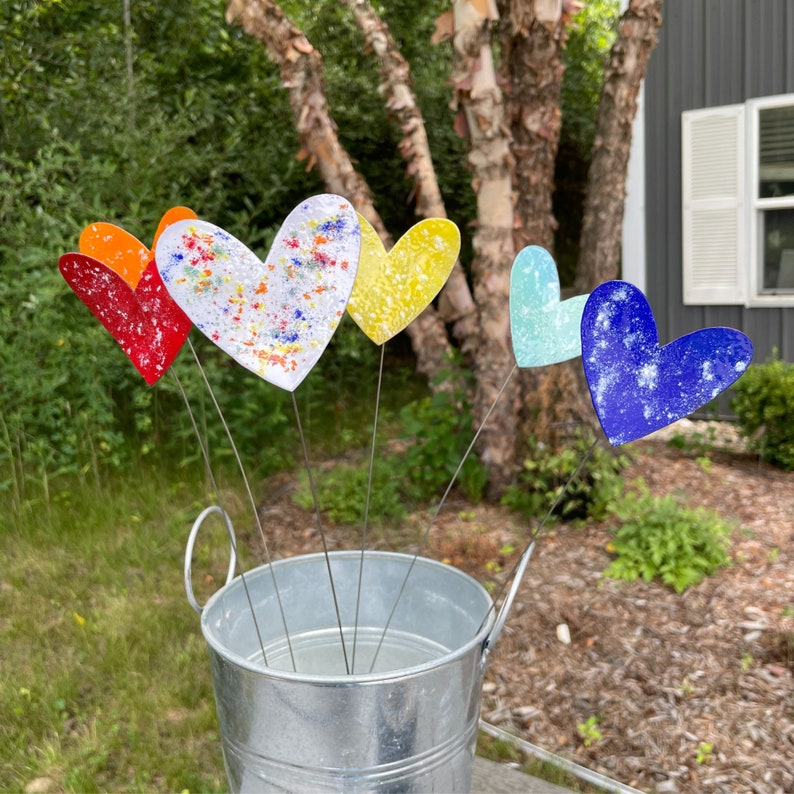 May include: A metal bucket contains a selection of heart-shaped garden stakes in various colours. The hearts are painted with speckled designs in red, orange, white, yellow, blue, and teal. The stakes are displayed outside in a garden setting.