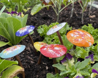 May include: A collection of colorful metal mushroom garden stakes with white speckles. The stakes are arranged in a garden setting with green plants and purple flowers.