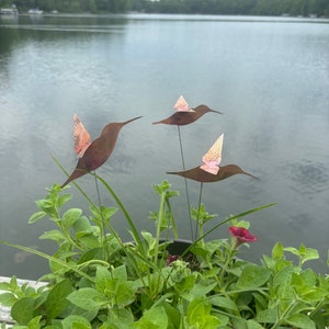 May include: Three copper hummingbird sculptures with pink accents are placed in a pot of green plants. The sculptures are on metal stakes and are positioned in a way that makes them look like they are flying.