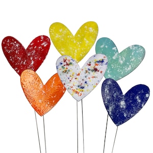May include: A selection of heart-shaped garden stakes in a variety of colours. The hearts are red, yellow, orange, white, blue, and teal. Each heart has a unique texture and design, with some featuring speckled patterns. The stakes are made of metal.