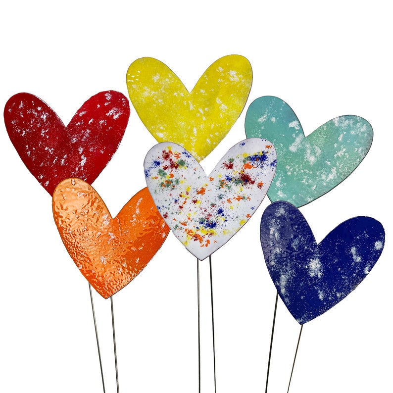 May include: A selection of heart-shaped garden stakes in a variety of colours. The hearts are red, yellow, orange, white, blue, and teal. Each heart has a unique texture and design, with some featuring speckled patterns. The stakes are made of metal.