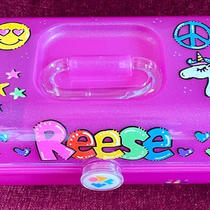 May include: A bright pink glittery plastic storage box with a clear handle and latch. The lid features a yellow smiley face, a blue peace sign, and a unicorn graphic. The name Reese is printed in colorful bubble letters, surrounded by small hearts and stars.