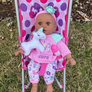 May include: A baby doll with dark skin, wearing a pink outfit and floral pants, sits in a pink stroller. The doll has a headband with a mint green bow and holds a white unicorn plush. The doll's shirt says "cora".