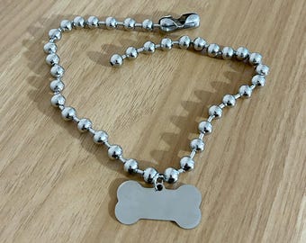 May include: A silver-colored dog tag in the shape of a bone, attached to a ball chain necklace. The tag is blank, ready for engraving. The chain has a clasp for easy attachment.