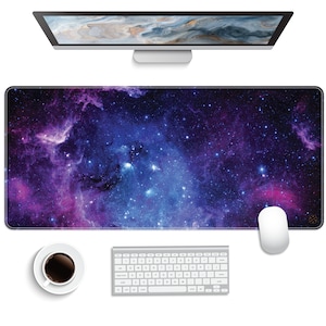 May include: A large purple and blue galaxy-themed mousepad with a white computer mouse on the right side. The mousepad is on a white surface with a white keyboard and a cup of coffee.