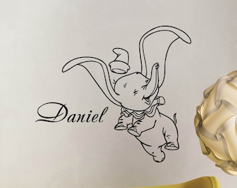 Puede incluir: Pegatina de pared de línea de arte en blanco y negro de Dumbo el elefante con un sombrero, volando con las orejas extendidas. El nombre "Daniel" está escrito en una fuente cursiva debajo del elefante.