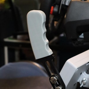 May include: A black metal racing handbrake with a gray plastic handle. The handbrake has a series of holes along the side and a textured grip.