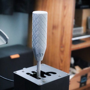 Op de afbeelding: Een zwarte metalen schakelbasis met een grijze, ruitvormige schakelknop. De knop heeft de tekst "eo" erop gedrukt.