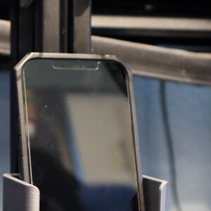 May include: A silver-colored phone holder with a black smartphone inside. The holder is mounted on a black vertical support, with a racing pedal visible in the foreground. The phone screen is black.