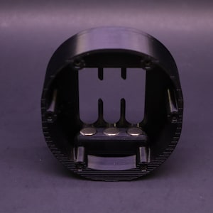 Pode incluir: Uma base circular de pl&aacute;stico preto com uma moldura retangular elevada e aberta no centro. A moldura tem tr&ecirc;s ranhuras verticais e uma fila de quatro pequenos &iacute;m&atilde;s redondos na parte inferior.