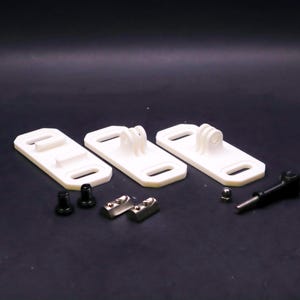 May include: A collection of white plastic camera mounts with various hardware components. The mounts have a rectangular base with a curved slot and a raised platform for attaching a camera. Additional hardware includes black screws and silver metal pieces.