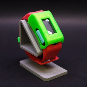 Peut inclure: Une montre connectée imprimée en 3D de couleur verte et rouge avec un petit écran et un bouton sur le côté, posée sur un support imprimé en 3D gris.