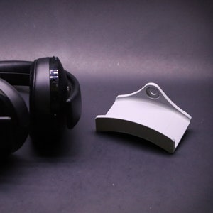 May include: A black pair of over-ear headphones with a white stand. The headphones are lying on a dark surface.