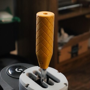 May include: A yellow gear shift knob with a diamond pattern, fitted to a white and grey gear shift assembly. The knob is cylindrical with a hole at the top. The assembly is part of a gaming setup.