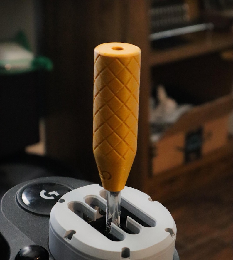 May include: A yellow gear shift knob with a diamond pattern, fitted to a white and grey gear shift assembly. The knob is cylindrical with a hole at the top. The assembly is part of a gaming setup.
