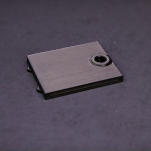May include: A small, rectangular, black metal object with a circular hole on one side. The object has a matte finish and appears to be a component or part of a larger mechanism. The item is set against a dark background.