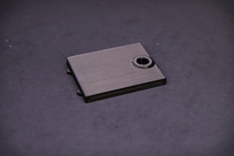 May include: A small, rectangular, black metal object with a circular hole on one side. The object has a matte finish and appears to be a component or part of a larger mechanism. The item is set against a dark background.